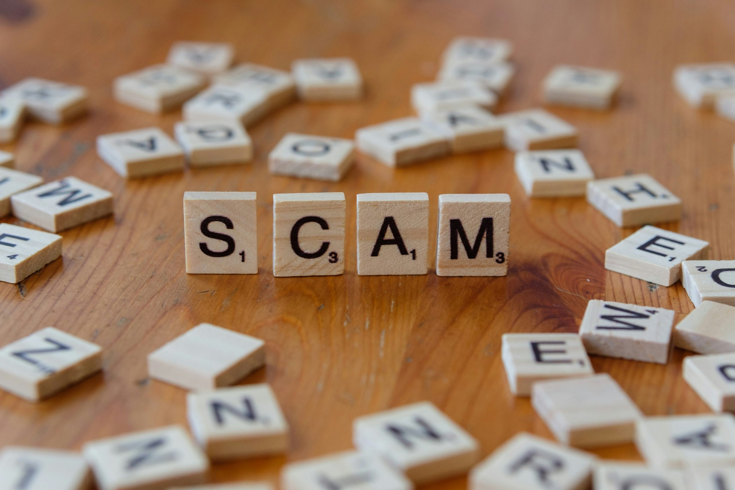 Scrabble tiles scattered on a table top. Four tiles spell "SCAM".