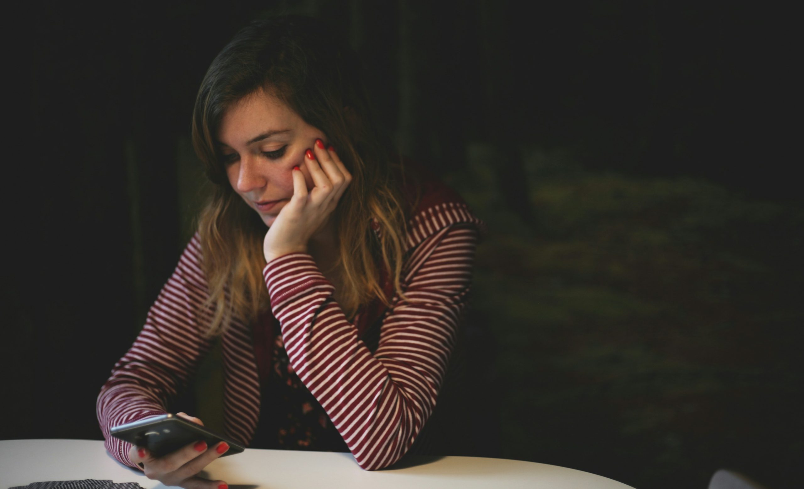 A woman leaning with her elbows on a table looking down at a cellphone with a worried expression, in a dark room