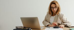 woman using a calculator at her desk which has a laptop and paperwork on it